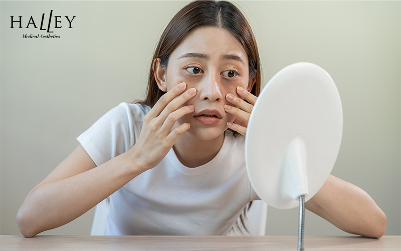 Person in white t-shirt examining face in round mirror with both hands in soft-lit setting.