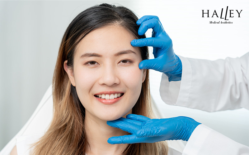 Smiling woman receiving facial assessment from gloved medical professional in clinical aesthetic setting.