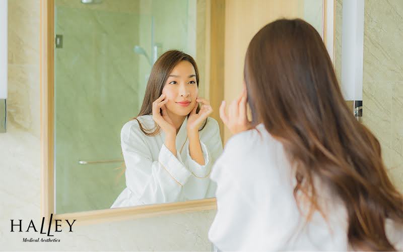 Person in white robe touching face while looking in bathroom mirror, suggesting self-care in modern setting.